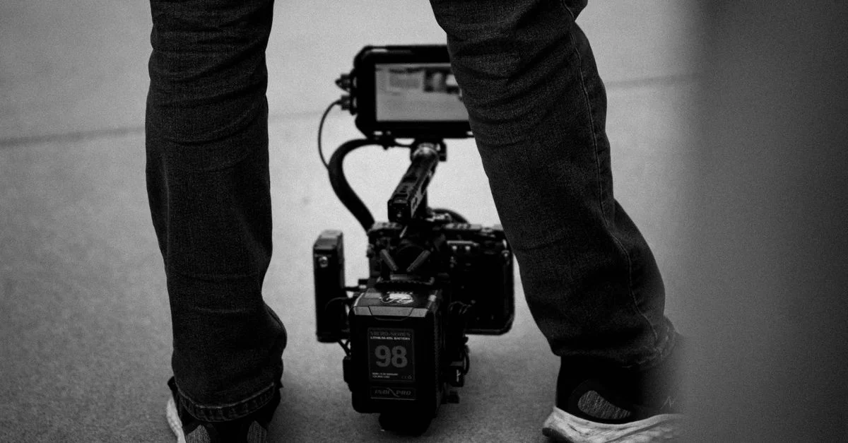 Photo en noir et blanc d'un cinéaste debout avec une caméra professionnelle posée sur le sol.